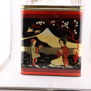 May include: A vintage tin box with a black background and a red, white, and black design. The box features a mountain scene with two figures in traditional Japanese clothing. The text on the box reads "Japan Tea".