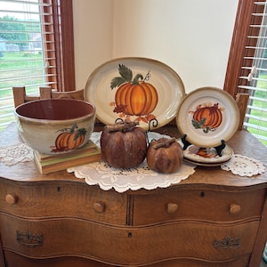 May include: A ceramic dinnerware set featuring a large oval platter, a deep bowl, and two plates, all decorated with painted orange pumpkins and autumn leaves. Two textured, brown, spherical pumpkin-shaped decorative items sit in the center.