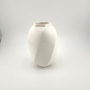 May include: A white ceramic vase with a geometric design. The vase has a hexagonal top and bottom, with a curved, spiraling shape. The vase is a solid, glossy white color.