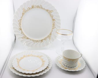 May include: A set of white porcelain dinnerware with a gold floral pattern. The set includes a dinner plate, a salad plate, a teacup, a saucer, and a wine glass with a gold rim.