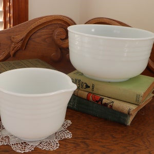 May include: Two white mixing bowls, one with a spout, on a stack of vintage books. The smaller bowl is on a white doily.