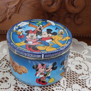 May include: A blue and white metal cookie tin featuring Disney characters Mickey Mouse, Minnie Mouse, Donald Duck, Goofy, and Pluto. The tin has the text "Fancy Fun Cookies - 300z (2 LBS) NET WT 32 OZ (2 LBS) - FANCY FUN COOKIES - 300z (2 LBS) NET" printed on the side.