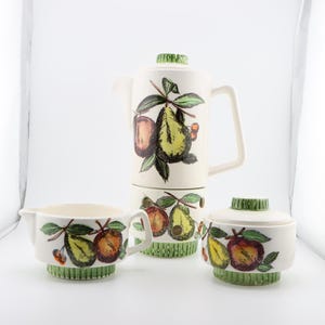 May include: A white ceramic tea set with a green and brown fruit design. The set includes a teapot, creamer, and sugar bowl.
