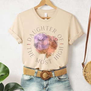 May include: A beige t-shirt with a graphic design of a woman's face with a butterfly on her shoulder. The text "Daughter of the Most High" is written in a circle around the woman's face.