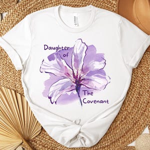 May include: White t-shirt featuring a watercolor-style purple flower design with the text "Daughter of The Covenant". The shirt has short sleeves and a crew neck.