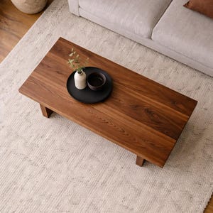 Peut inclure: Table basse rectangulaire en bois brun fonc&eacute; avec un fini grain de bois naturel. Un plateau noir contient un petit vase blanc avec de la verdure et un bol brun fonc&eacute;. La table est pos&eacute;e sur un tapis de couleur claire.