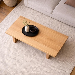 Peut inclure: Table basse rectangulaire en bois clair avec une finition naturelle. Un plateau noir supporte un petit vase blanc avec de la verdure et un bol marron fonc&eacute;. La table est pos&eacute;e sur un tapis clair.