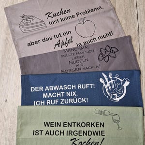 Könnte beinhalten: Ein Stapel Küchentücher in verschiedenen Farben, jeweils mit einem humorvollen deutschen Satz in Schwarz bedruckt. Die Tücher umfassen ein graues mit Kuchen und Apfel, ein dunkelgraues mit Nudeln, ein blaues mit Küchenutensilien und ein grünes mit einer Weinflasche.