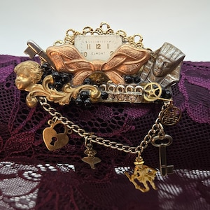May include: A decorative brooch featuring a central Elmont watch face, a copper-toned bow, a gold-toned cherub, and a silver-toned pharaoh bust. It includes black beads, clear rhinestones, and a gold-toned chain with charms: a heart lock, a dancer, a key, and a figure.