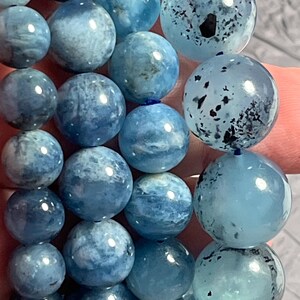 Op de afbeelding: Close-up van een armband gemaakt van ronde, lichtblauwe aquamarijn kralen. De kralen hebben een doorschijnende kwaliteit met verschillende blauwtinten en enkele donkere insluitsels. De armband is geregen op een blauw koord.