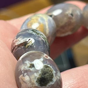 Op de afbeelding: Een armband bestaande uit gladde, ronde, gepolijste kralen in tinten van gedempt lavendel en grijs. Elke kraal heeft unieke, natuurlijke insluitingen met vlekken van donkergroene, witte en bruine mineraalafzettingen op het oppervlak.