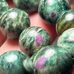 Peut inclure: Une collection de perles sphériques polies présentant un motif marbré vert et blanc avec des inclusions distinctes et vibrantes rose magenta. Chaque perle a une finition lisse et brillante qui reflète la lumière, soulignant la texture.