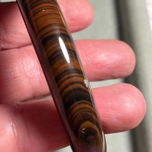Op de afbeelding: Een gepolijste, cilindrische edelsteen met een tijgeroogpatroon. De steen heeft bruine en zwarte banden, waardoor een gestreept effect ontstaat. De edelsteen wordt in een hand gehouden en toont zijn gladde, glanzende oppervlak.
