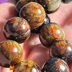 Può includere: Un bracciale composto da perline sferiche levigate con un motivo vorticoso nei toni del marrone scuro, arancione dorato e blu scuro. La superficie liscia e riflettente di ogni perlina mostra inclusioni minerali naturali e riflessi.