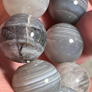 Peut inclure: Gros plan sur un bracelet composé de grosses perles rondes. Les perles sont principalement grises avec des veines blanches et grises plus foncées, créant un effet marbré. Les pierres polies ont une finition brillante, reflétant la lumière. C'est un bijou.