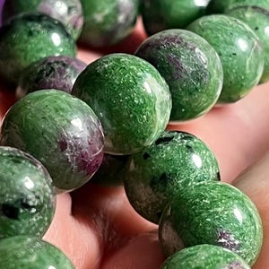 Pode incluir: Um colar de contas esféricas polidas de zoisite com inclusões de rubi. As contas apresentam uma cor base verde vibrante com texturas verde-escuras manchadas e manchas irregulares distintas de magenta profundo e roxo por toda a pedra.