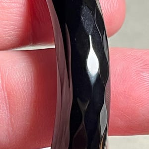 Peut inclure: Un bracelet jonc noir brillant présentant une texture martelée distincte sur toute sa surface. La bande incurvée et solide possède une finition réfléchissante qui capte la lumière, créant des reflets brillants le long du motif facetté.