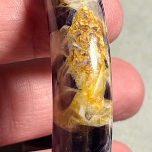 Peut inclure: Gros plan sur un bracelet poli et translucide. Le bracelet présente un mélange de teintes violet foncé, crème et jaune doré, créant un effet marbré. Les couleurs sont tourbillonnées et superposées, donnant au bracelet un aspect naturel unique. Le bracelet est un bijou.