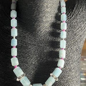 Puede incluir: Un collar con cuentas rectangulares de color azul claro intercaladas con pequeñas cuentas rosas. El collar también incluye detalles de cristal transparente y cuentas de metal plateado. Se muestra sobre un maniquí negro.