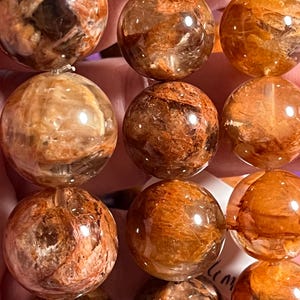 Op de afbeelding: Een streng van ronde, gepolijste kralen gemaakt van helder kwarts met opvallende, dichte, roodbruine en goudoranje minerale insluitingen. De kralen zijn in verticale rijen gerangschikt en hebben een glad, reflecterend oppervlak.
