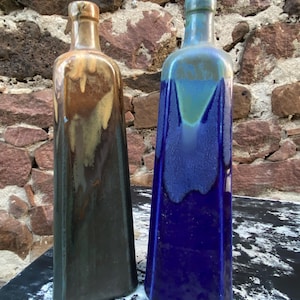 May include: Two ceramic bottles with a rectangular shape. One bottle is glazed in shades of brown and gold, the other in blue and teal. The bottles have a rustic, handmade appearance and are displayed against a stone wall.