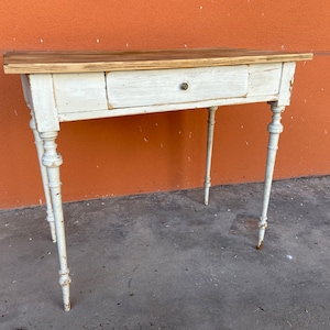 Puede incluir: Mesa de madera pintada de blanco con un cajón y patas torneadas. La superficie de la mesa es de madera natural.
