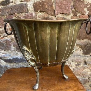 Peut inclure: Un grand pot en laiton antique avec un design cannelé et trois pieds ornés. Le pot a deux poignées en anneau et des accents décoratifs. Le laiton a une patine chaude et vieillie. Le pot est exposé sur une table en bois contre un mur en pierre.
