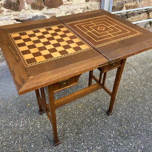 May include: A wooden game table with a fold-down top that features a checkerboard and a geometric pattern. The table has a simple, Arts and Crafts style with a dark wood finish and brass accents.
