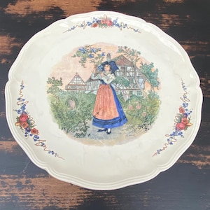 Puede incluir: Plato decorativo color crema con borde festoneado. El centro presenta una escena pintada de una mujer con vestimenta tradicional frente a una casa. Diseños florales y de bayas adornan el borde.