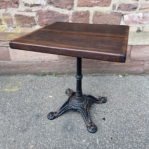 May include: A dark brown wooden table top with a black cast iron base. The table has a square top and four legs with intricate details.