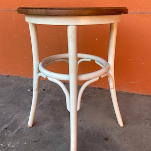 May include: A small, round, white wooden stool with a dark brown tabletop. The stool has a curved design with a circular footrest and four legs. The stool is approximately 18 inches tall.