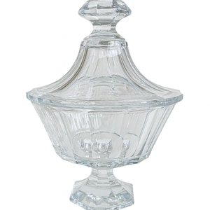 May include: A clear, faceted glass candy dish with a lid. The dish has a pedestal base and a flared bowl shape. The lid has a diamond-shaped finial. The glass has a ribbed design. A decorative piece for serving or display.