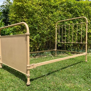 May include: A vintage metal bed frame in a light beige color. The bed frame has a headboard with a flat panel and a simple design. The frame is made of metal and has a slightly distressed finish.