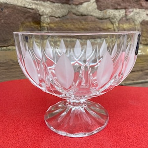 May include: A clear glass bowl with a frosted design of leaves. The bowl has a pedestal base and is sitting on a red surface.