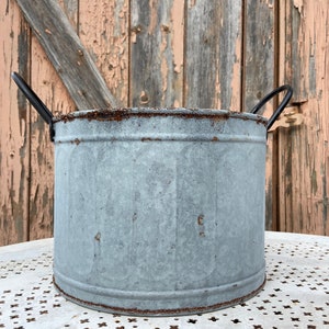 Może przedstawiać: Galwanizowana metalowa doniczka z dwoma czarnymi metalowymi uchwytami. Doniczka ma rustykalny, zużyty wygląd i stoi na białym stole z wzorzystą powierzchnią.