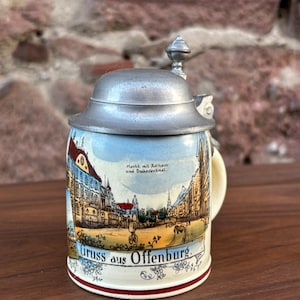 May include: A ceramic beer stein with a metal lid. The stein features a detailed illustration of a European town scene, with the words "Gruss aus Offenburg" at the bottom. The handle is cream-colored, and the base has a red stripe.