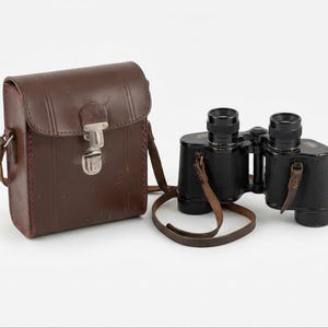 Könnte beinhalten: Ein schwarzes Fernglas mit strukturierter Oberfläche und einem dünnen braunen Leder-Umhängeriemen, neben einer rechteckigen braunen Ledertasche mit einem silberfarbenen Metallverschluss und passenden seitlichen Lederriemen.