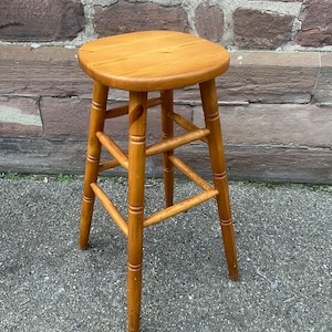May include: A wooden stool with a natural finish and a rounded top. The stool has four legs and a simple design.