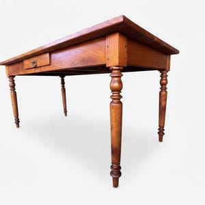 Puede incluir: Una mesa rectangular de madera con un acabado marrón cálido, que presenta una superficie superior plana, cuatro patas torneadas con detalles decorativos y un cajón único situado en el faldón frontal con un pequeño tirador circular de metal.
