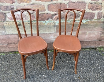 May include: Two brown wooden chairs with a simple design. The chairs have a curved back and a slatted seat.