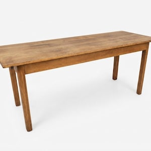 Puede incluir: Una mesa rectangular de madera con un acabado marrón natural, que presenta una superficie de tablero lisa y plana junto con cuatro patas rectas y robustas. La construcción muestra una textura de veta de madera visible en todo el mueble.