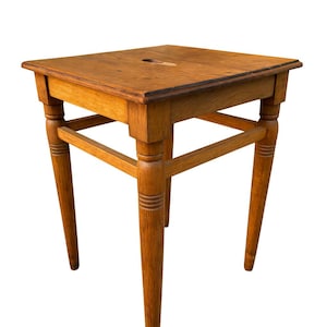 May include: A small, square wooden stool with a natural finish. The stool has a square top with a small hole and four tapered legs. The legs are connected by horizontal supports. The stool is approximately 18 inches tall.