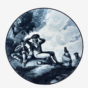 May include: A circular decorative plate featuring a monochromatic blue and white painted scene of four figures in a landscape. One central figure sits adjusting a hat, while others are seated nearby under a tree against a cloudy sky background.