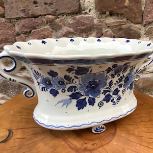 May include: White ceramic planter with blue floral designs and handles. The planter has a scalloped rim and is decorated with blue flowers and leaves. It sits on small feet and has a blue border around the base and rim.