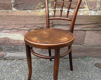 May include: A vintage wooden chair with a round seat and curved back. The chair has a dark brown finish, with a backrest featuring vertical spindles. The legs are slightly curved, and the chair appears to be made of solid wood.