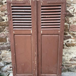 Puede incluir: Un par de contraventanas de madera marrón con paneles de persiana y herrajes metálicos. Las contraventanas están cerradas y tienen un aspecto desgastado.