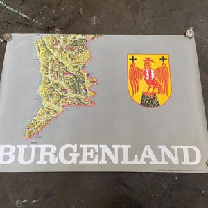 May include: A vintage poster featuring a map of Burgenland, Austria, with a coat of arms depicting a red eagle with a yellow shield and two crosses. The poster has a gray background and the text "Burgenland" in white.