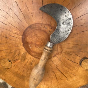 May include: A vintage sickle with a curved, steel blade and a wooden handle. The blade shows signs of wear, and the handle has a smooth, aged appearance. The sickle is resting on a wooden surface.