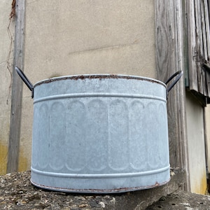 May include: A grey metal planter with black handles. The planter has a slightly distressed look and is sitting on a stone surface.
