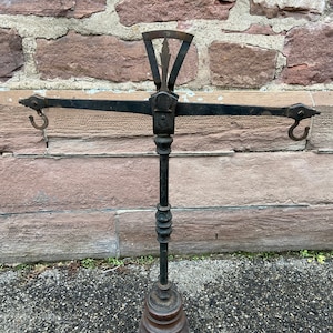Peut inclure: Une balance à suspendre ancienne en métal noir avec une base en bois. La balance a un bras en métal avec deux crochets à chaque extrémité et un cadran circulaire avec des marques.